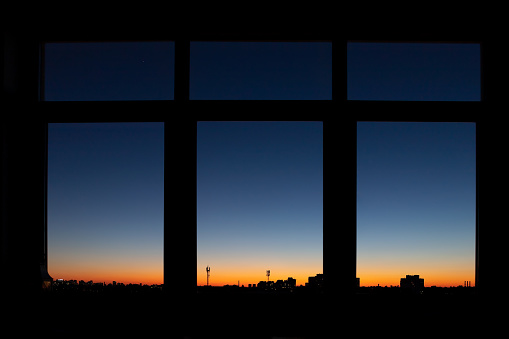 Through triple window POV with scenic city downtown cityscape black building silhouettes against fiery orange to blue dramatic sunset or sunrise on early morning time. Metropolis panoramic background.