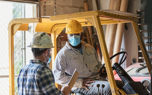 Two multi-ethnic mature men in their 40s working in a warehouse, wearing hard hats and safety goggles, conversing. The forklift operator is an African-American man. His coworker is standing next to the forklift, holding a clipboard. They are wearing protective face masks, working during the covid-19 pandemic, taking precautions to prevent to spread of coronavirus.