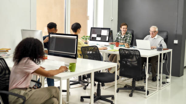 istock Full length shot of aged man, senior intern typing, using laptop while sitting at desk, working together with other young employees in modern office 1283406785
