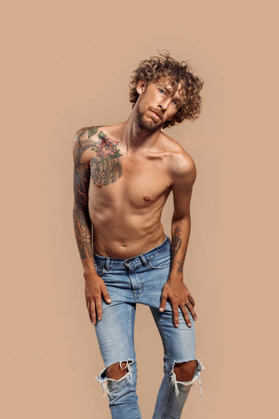 freestyle. uomo con i capelli ricci e la parte superiore nuda del corpo in jeans in piedi isolati su beige in posa pensieroso - stubble men tattoo sensuality foto e immagini stock