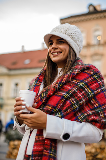 Low angle view of a smiley brunette in white winter clothes and a chekered scarf drinking a hot beverage in styrofoam cup in a public area.
