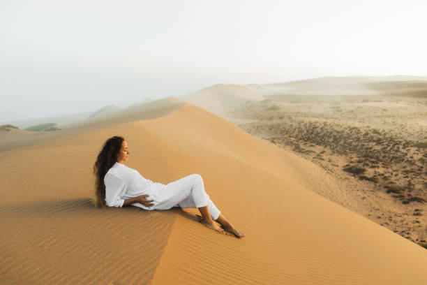 モロッコサハラ砂漠の砂丘の上でリラックス白い服を着た美しいブルネットの女性。日の出前の朝の霧。自然との調和 - desert landscape morocco sand dune ストックフォトと画像