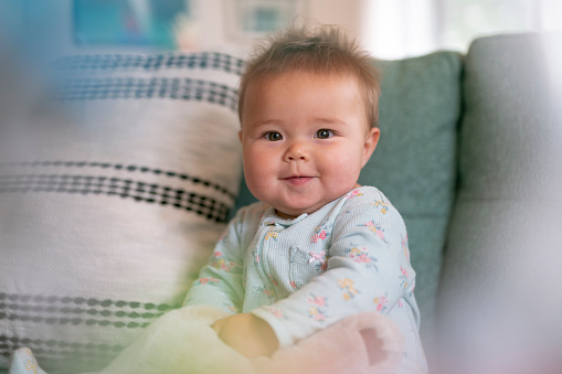 Adorable mixed race baby wearing pajamas smiles. directly at the camera with pride as she sits up by herself on the couch in the living room of her home.