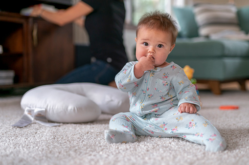 Cute mixed race baby girl wearing pajamas sitting up by herself in the living room of her home. The child is looking directly at the camera as her mom organizes toys in the background.
