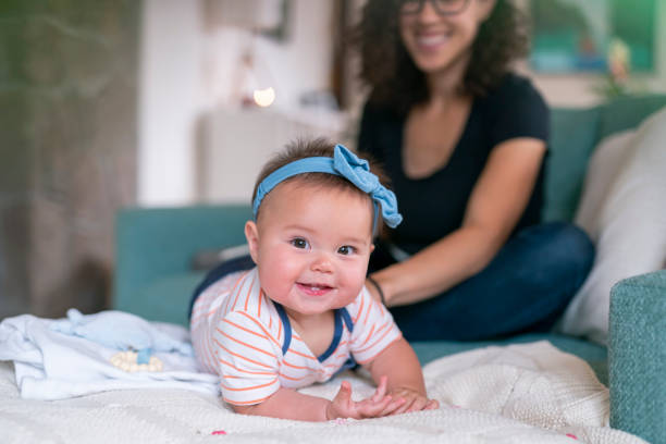 A cute mixed race baby girl smiles directly at the camera while lying on her tummy on the couch at home. The baby's mother is in the background sitting on the couch and smiling with pride at her growing baby.
