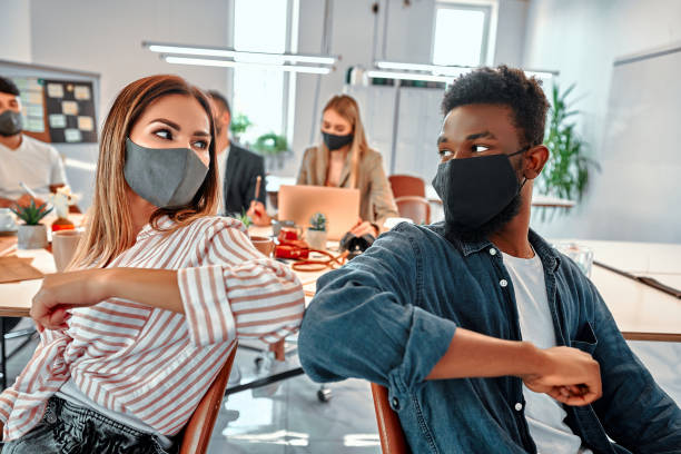 Two colleagues in medical masks avoid a handshake when meeting in the office greeting with bumping elbows during coronavirus COVID-19 epidemic in office