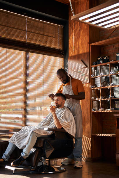 Bearded male barber cutting client hair with scissors Handsome young man sitting in barber chair and holding glass of drink while getting haircut in barbershop black barber stock pictures, royalty-free photos & images