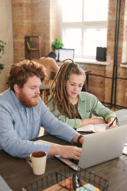 istock Busy young red-bearded man typing on laptop while preparing report with colleague in coworking space 1283436251
