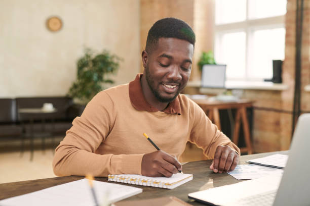 istock Content handsome young Black man in sweater sitting at table and making notes in sketchpad while summarizing information of online course 1283436937