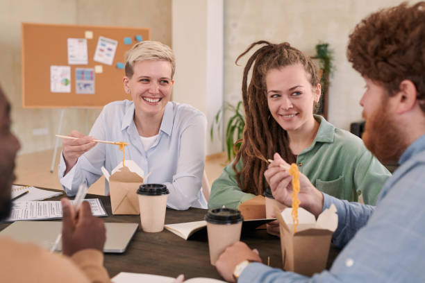 istock Cheerful young coworkers sitting at table and chatting with each other while eating restaurant dishes in office 1283436960