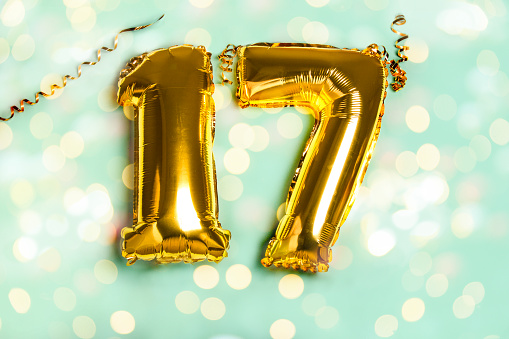 Decoration for seventeen birthday party. Golden balloon in form of 17 number on blue background. Sparkles wallpaper decor