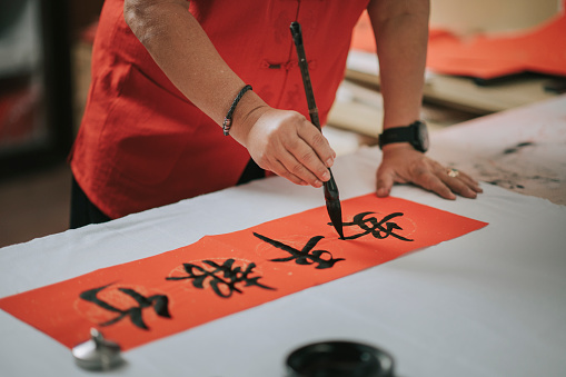 hand of asian chinese male practising chinese caligraphy for coming chinese new year celebration home decoration purpose with prosperity and good wording by writing it on a red piece of paper