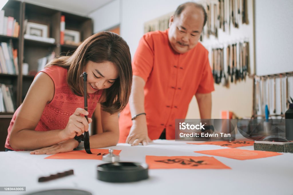 An asian chinese male practising chinese caligraphy for coming chinese new year celebration home decoration purpose with prosperity and good wording by writing it on a red piece of paper and teaching his daugther Chinese Culture Stock Photo