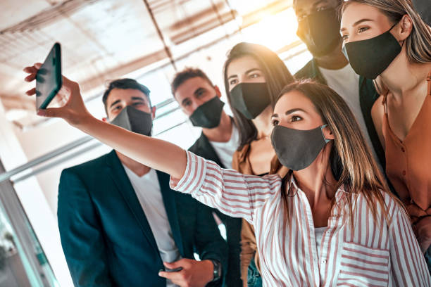 jóvenes positivos complacidos colegas de negocios en el interior en la oficina tomar un selfie usando máscaras debido al coronavirus. - break office 30s 20s fotografías e imágenes de stock