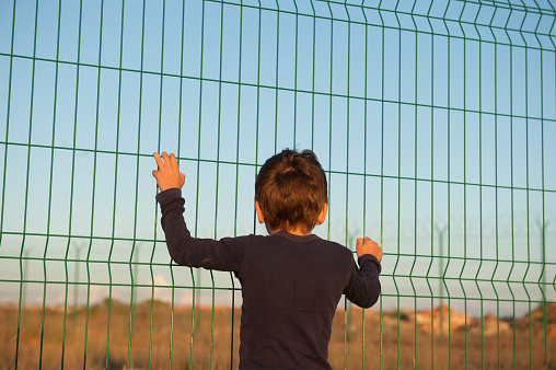 hope refugee concept one little prisoner boy in black shirt holding fence on Mexico state border in desert on sunset in displaced persons camp