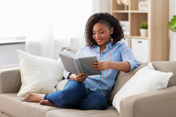 happy african american woman reading book at home leisure, literature and people concept - smiling african american woman reading book at home reading stock pictures, royalty-free photos & images