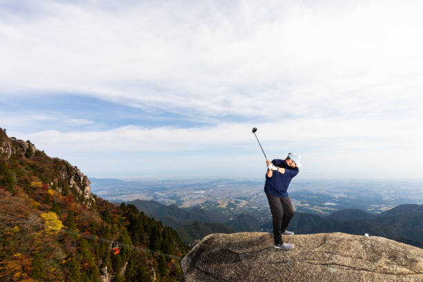 A male Golfer teeing off high up on a mountain in a precarious spot with a city visible way off in the distance bvelow. There is copy space around the image for text.