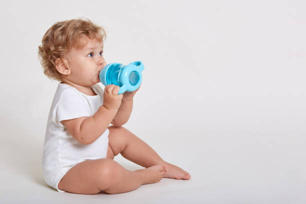 하얀 벽에 앉아 있는 동안 병에서 물을 마시는 귀여운 유아의 초상화, 바디 슈트를 입고, 곱슬 머리가 목마른 한 살 짜리 아이, 유아의 측면 보기. - baby cup 뉴스 사진 이미지