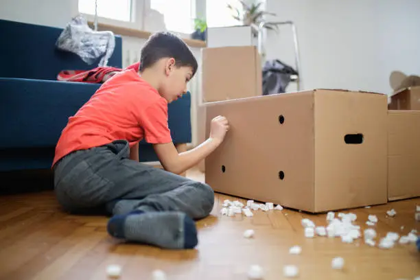 Photo of Boy sitting on floor and pushing packing peanut through a hole on cardboard box