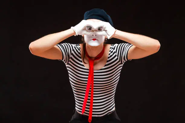 Portrait of male mime artist performing, isolated on black background. Man folds his hands as if looking through binoculars.