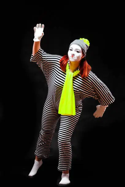 Portrait of female mime artist, isolated on black background. Young woman in striped suit and bright yellow scarf and hat is pretending crawling.