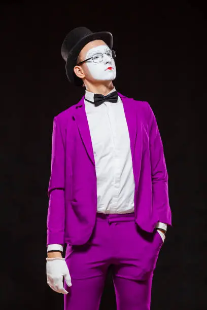 Portrait of male mime artist, isolated on black background. Young man in purple suit is standing with risen finger. Symbol of attention, warning, nota bene.