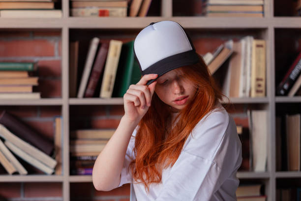 Portrait of a redhead woman in template blank t-shirt and cap with healthy freckled skin. Young caucasian model posing on bookshelf background. Copy space and mock up. Place for adverising Portrait of a redhead woman in template blank t-shirt and cap with healthy freckled skin.Young caucasian model posing on bookshelf background. Copy space and mock up. Place for adverising woman wearing baseball cap stock pictures, royalty-free photos & images