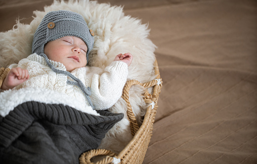 Portrait of a sleeping baby in a wicker cradle in a warm knitted hat under a warm blanket with a toy in the handle. Close up.