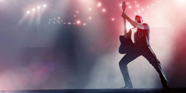 Rock Star Holding Up Guitar On Stage A rock star dressed in short sleeved shirt and leather trousers, slightly squatting with legs apart, shouts and holds up a generic electric guitar on stage. The musician is alone on stage which is lit by many spotlights. With copy space. rock musician photos stock pictures, royalty-free photos & images