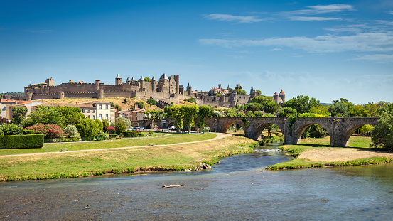 Carcasonne cityscape panorama view over aude river in summer under blue sunny sky. Medieval fortified stone wall on hill top, Pont Vieux old stone bridge crossing the Aude river on the left. Carcasonne, Languedoc Rousillon, South France, France, Europe.