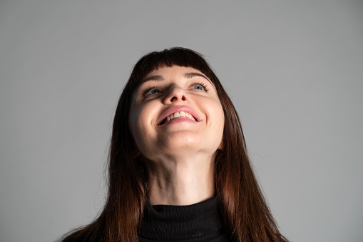 Close up studio portrait of a pretty brunette woman, wearing folded black polo-neck sweater, laughing, looking to the side, against a plain grey background