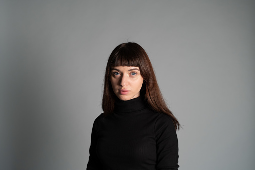 Studio portrait of a pretty brunette woman, wearing folded black polo-neck sweater, looking at camera, against a plain grey background