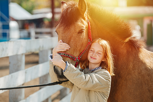 Happy preteen ginger girl holding horse's snout with both hands, touching horse's head with hers. Her eyes closed. Against sunlight.