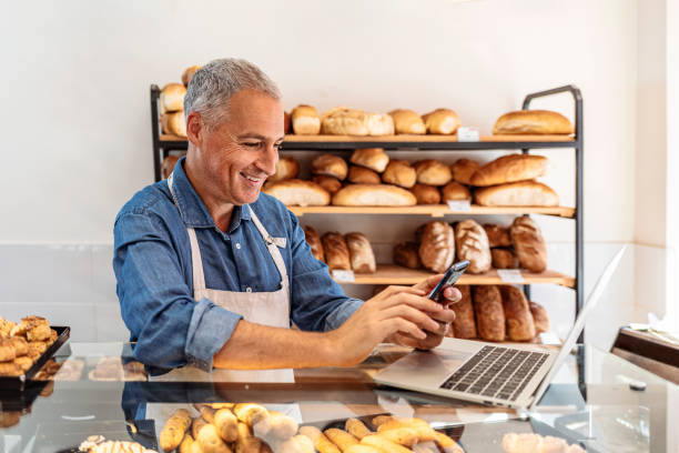 Mature man using laptop for online business order Cropped shot of a handsome mature businessman working in his local internet cafe or bakery shop. baker occupation stock pictures, royalty-free photos & images