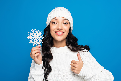 happy woman in knitted hat holding decorative snowflake and showing thumb up isolated on blue