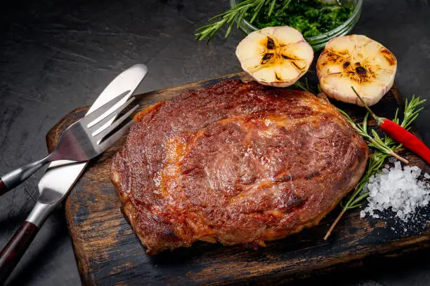 Well done grilled ribeye beef steak on a wooden board on dark stone background, near fork, knife and seasoning.