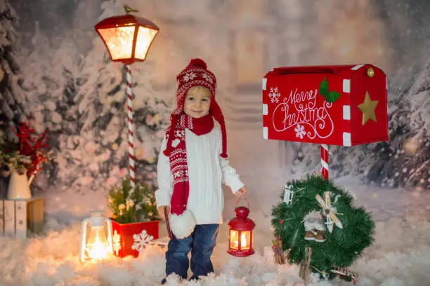 Photo of Little child, toddler boy, sending letter to santa in christmas mailbox, christmas decoration around him, outdoor shot