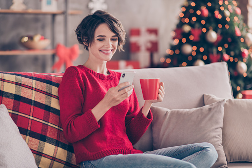 Photo of positive lady sit cozy couch use cellphone hold beverage mug in house indoors with christmas x-mas ornament.