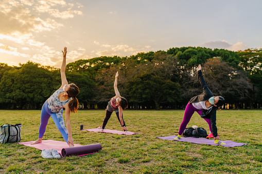 A group of women are practicing yoga in a public park while keeping social distancing and covering their faces with protective face masks.