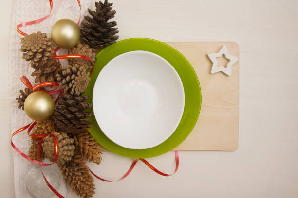 istock Empty flat on a white wooden table with New Year decorations. Pine cones, Christmas balls, ribbon and bokeh on a white empty plate with a wooden stand. Christmas food concept, top view. Copyspace 1283491867