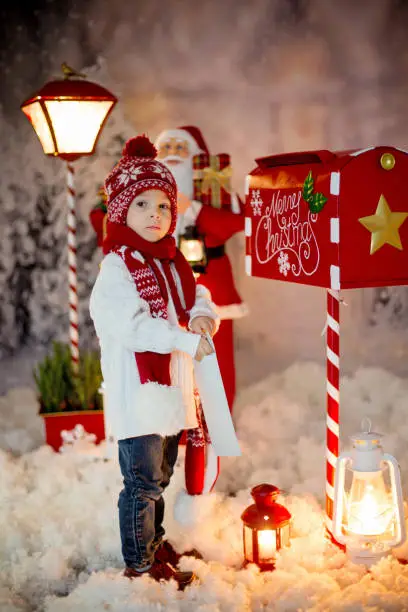 Photo of Little child, toddler boy, sending letter to santa in christmas mailbox, christmas decoration around him, outdoor snow shot