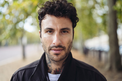 Headshot of male with curly black hair, mustache and beard stubble, wearing casual clothing and looking at camera with serious expression.