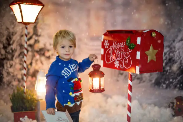 Photo of Little child, toddler boy, sending letter to santa in christmas mailbox, christmas decoration around him, outdoor snow shot