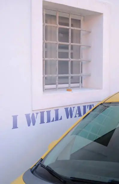 "I WILL WAIT" is written on a wall, behind a yellow parked car. *** The stencil was digitally added and a release is provided ***