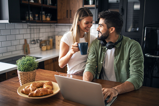 Two people in love smiling at each other. Man is typing on laptop and women is holding coffee happily