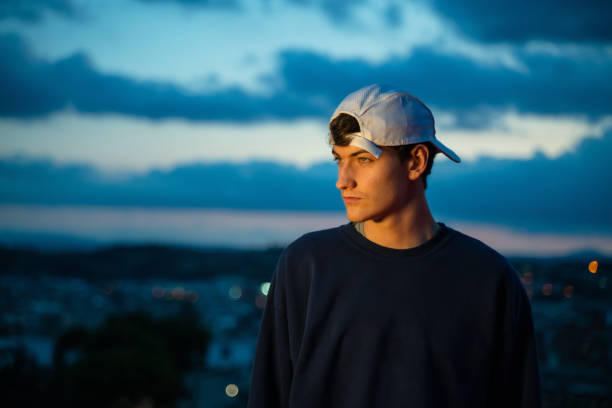 Cool young man by city lights at evening. He is wearing a dark blue sweater and a cap.