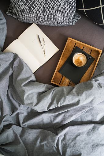 Breakfast in bed. Cup of coffee on the black notebook on the wooden tray and open book with lavender flowers on the bed with grey blanket. Flat lay.