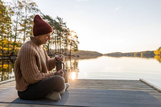 Autumn morning A woman sitting by a lake in a forest during autumn. She is drinking tea out of a travel mug and is wearing a knitted hat and sweater. swedish woman stock pictures, royalty-free photos & images
