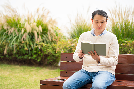 Asian man reading a book and sitting on a park bench.