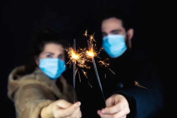 Photo of Young couple wearing protective face masks for Coronavirus and holding sparklers for New year's eve, 2021 and Covid-19 concept